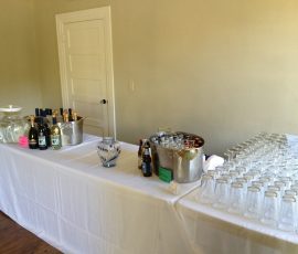 A table with bottled drinks, ice buckets, pitchers, and rows of empty glasses set up for a self-serve beverage station.