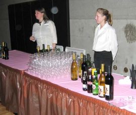 Two women stand behind a table with wine, beer, soda bottles, stacked glasses, and plastic cups at an event.