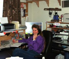 Woman in a purple shirt sits at a cluttered home office desk, talking on the phone and using a computer.