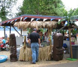 Outdoor tiki-themed food stand with thatched roof, tiki statues, and people gathered around on a sunny day.