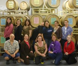 A group of people pose in front of stacked wine barrels in a winery or storage room.