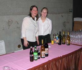 Two women stand behind a table with various bottles of drinks and glasses, ready to serve at an event.