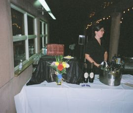 A woman stands behind a table with wine bottles, flowers, and glasses at an outdoor event or reception.