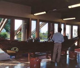A wooden canoe with painted designs is used as a bar inside a room with large windows; people are gathered nearby.