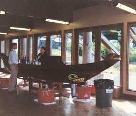 Two people stand near a wooden canoe-shaped counter inside a building with large windows and bright lighting.