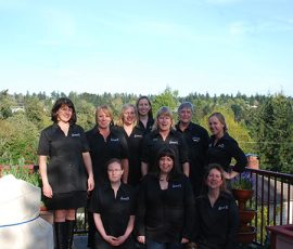 Ten women in matching black shirts pose together outdoors on a deck with trees and a blue sky in the background.