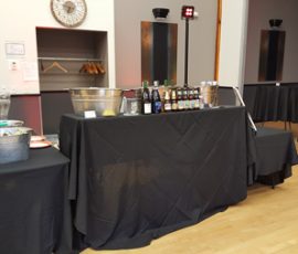 A black-draped bar setup with bottled drinks and ice buckets, in a room with empty coat racks and round tables.