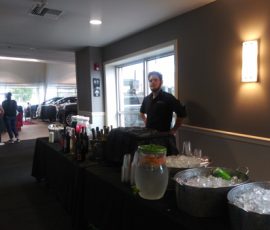 A man stands behind a beverage table with bottles, an ice bucket, and a water dispenser at an indoor event.