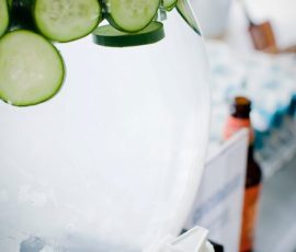 A clear beverage dispenser with sliced cucumbers floating in water and a white spigot at the bottom.