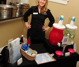A woman in black uniform stands behind a refreshment table with two large beverage dispensers and a basket of straws.
