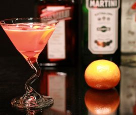 A pink cocktail with a lemon twist in a wavy glass, with an orange and liquor bottles in the background.