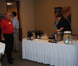 A woman arranges drinks on a table with wine, beer, and water as people gather nearby in a room.