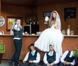 Bride in white dress sits on bar holding a beer, while three groomsmen nap beneath her and another gives a thumbs up.