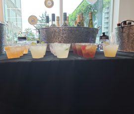 A row of assorted cocktails in plastic cups is displayed on a black tablecloth in front of bottles and ice buckets.
