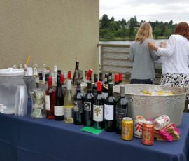 Table with assorted wine bottles, canned drinks, water dispensers, and two women talking by a railing overlooking water.