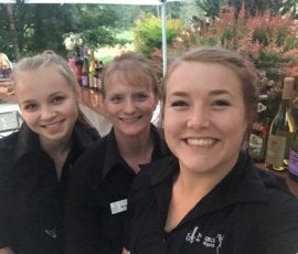 Three women in black shirts smile at the camera in front of a table with bottles of wine outdoors.