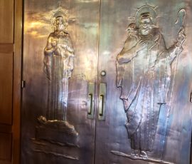 Metal doors with embossed religious figures and Latin inscriptions in a wood-paneled interior.