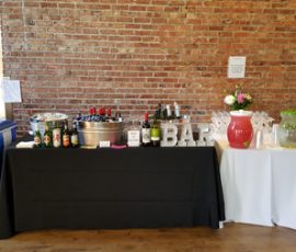 A bar setup with drinks, glassware, and flowers on tables against a brick wall, with recycling bins nearby.