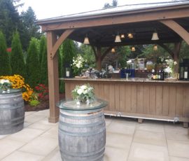 Outdoor wooden bar under a gazebo with floral arrangements and two barrel tables on a stone patio.