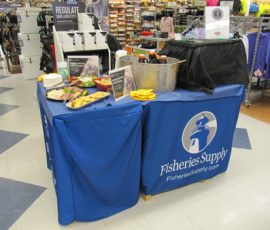 A promotional table for Fisheries Supply with refreshments and brochures, set up inside a retail store.