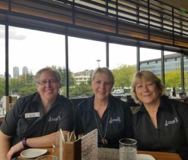 Three women in matching black shirts sit at a restaurant table with city buildings visible through large windows behind them.