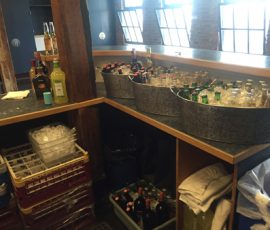 A bar counter with bottles of liquor, metal tubs of bottled drinks, an ice bin, and shelves with towels and glassware.