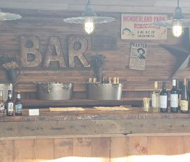 Rustic bar setup with metal tubs, bottles, drinks, and vintage signs on a wooden wall under hanging lights.