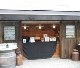Open wooden barn doors reveal a counter with a black tablecloth and lights inside, flanked by barrels and milk cans.