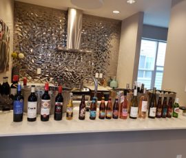 A variety of wine and beer bottles are lined up on a modern kitchen counter with a decorative backsplash.