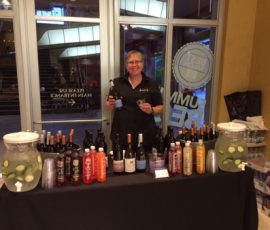 A person stands behind a table displaying bottles and drinks, holding a wine bottle and glass in a lobby area.