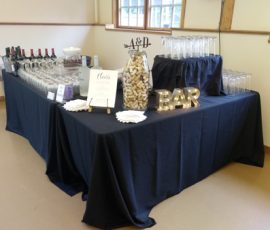 A bar table with wine bottles, glasses, a cork centerpiece, and a lit "BAR" sign set up for an event.