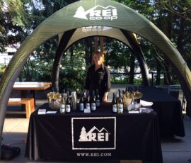 A person stands behind a table with REI branding, displaying bottles and ice buckets under a canopy outdoors.