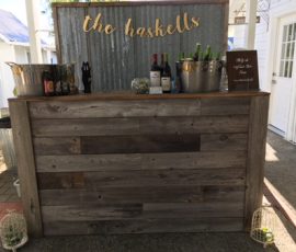 A rustic wooden bar with drinks, labeled "the haskells," set up outdoors with bottles and ice buckets on the counter.