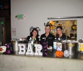 Three women stand behind a decorated bar with Halloween-themed items and large letters spelling "BAR.