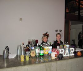 Two women stand behind a bar with various drinks; festive decorations and a lit "BAR" sign are on the counter.