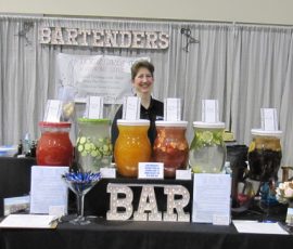 A woman stands behind a bar with five large drink dispensers filled with colorful beverages and fruit.