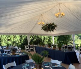 Outdoor tented event with round tables, navy tablecloths, white chairs, and a chandelier with greenery overhead.