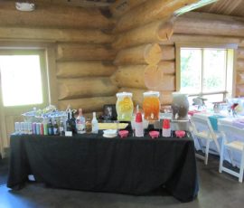 A drink station with bottled drinks, dispensers, cups, and mixers set up on a black tablecloth in a log cabin room.