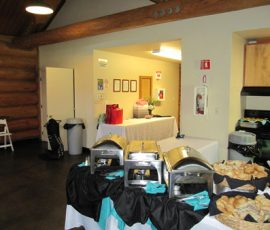 Buffet setup with covered chafing dishes, a basket of bread, and tables in a room with log walls and kitchen appliances.
