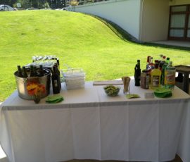 A white tablecloth-covered table with drinks, bottles, cups, napkins, and a small vase of flowers set up outdoors.
