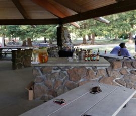 Stone pavilion with picnic tables, assorted drinks on a counter, and people standing and sitting outdoors in a park setting.