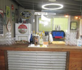 A bar counter with wine bottles, glasses, hand sanitizer, snacks, and signs under a circular ceiling light.