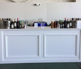 White bar counter with assorted bottles of alcohol and mixers arranged in ice buckets and rows on top.