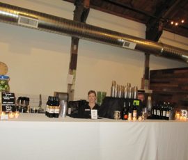 A bartender stands behind a white-clothed bar with bottles, mixers, and candles in a rustic indoor setting.
