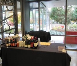 Table with wine bottles, soda cans, plastic cups, and snacks set up by large windows overlooking a garden area.
