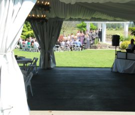 View from inside a tent looking out at an outdoor wedding ceremony with guests seated on the lawn.