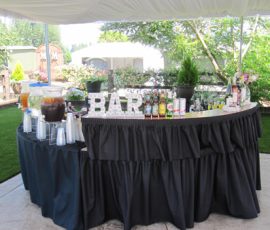 Outdoor bar setup with black tablecloths, drink dispensers, bottles, cups, and a "BAR" sign under a canopy.