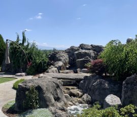 A landscaped garden with large rocks, a small stream, greenery, and a clear blue sky overhead.