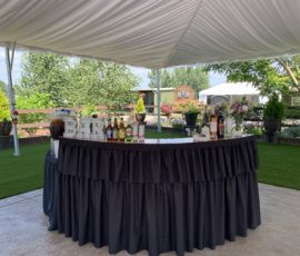 Outdoor bar setup with assorted bottles and drink dispensers under a covered canopy, surrounded by greenery.