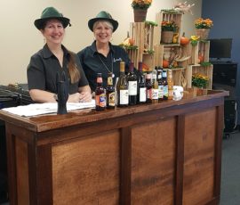 Two women in hats stand behind a bar with various drinks on display; shelves with pumpkins and flowers in the background.